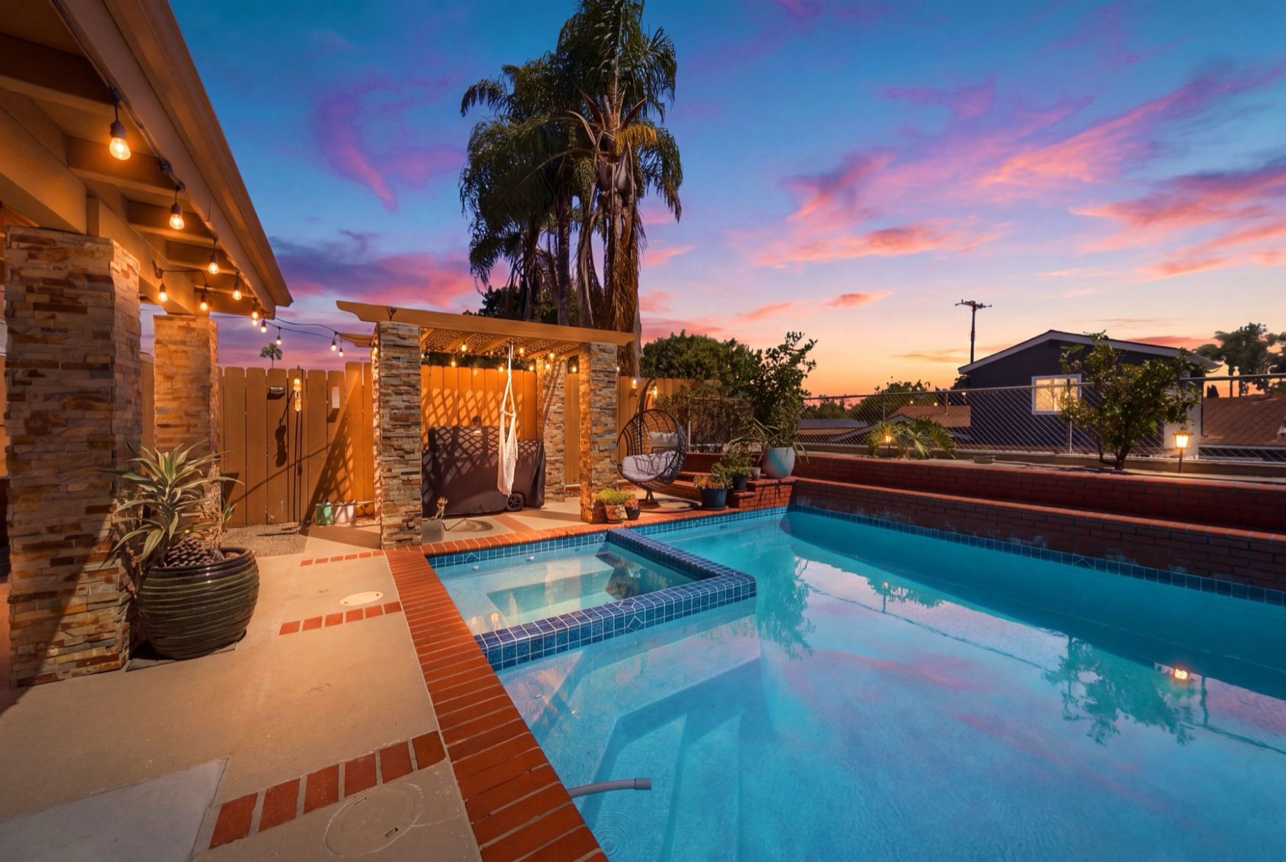 Backyard pool and spa at sunset with string lights and a stone pergola